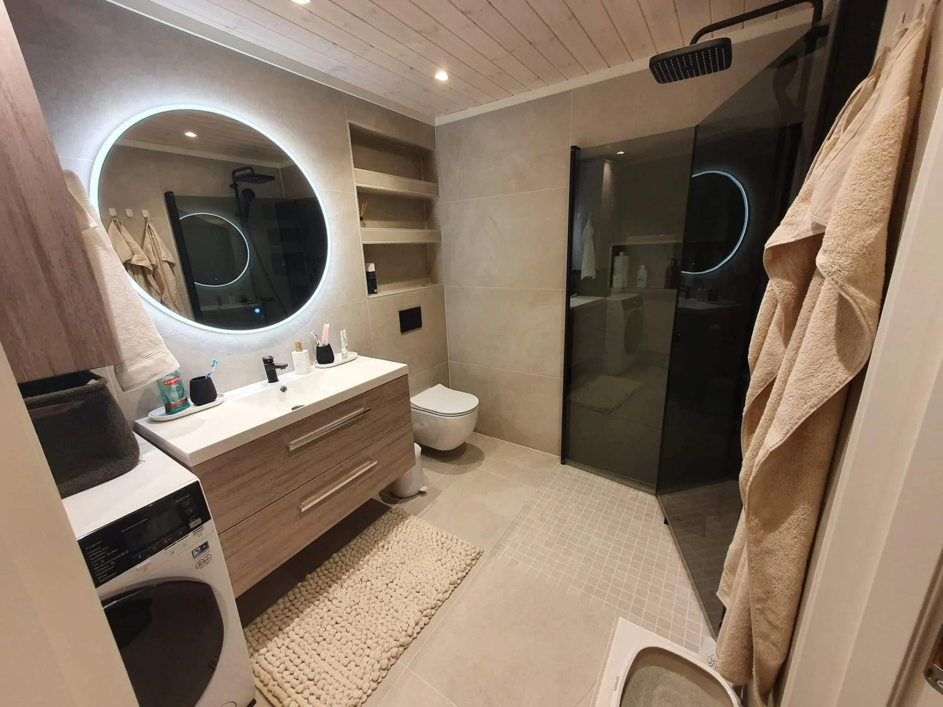A finished bathroom with a round lit mirror, a walk-in shower and a washing machine