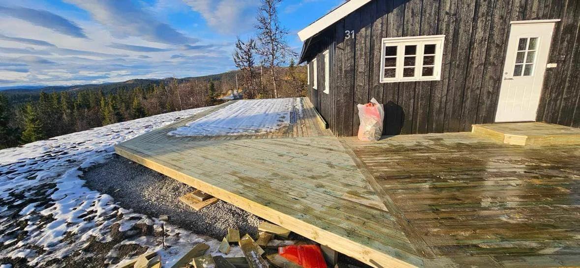 New terrace boards laid in front of a dark-stained cabin, snow on the hillside